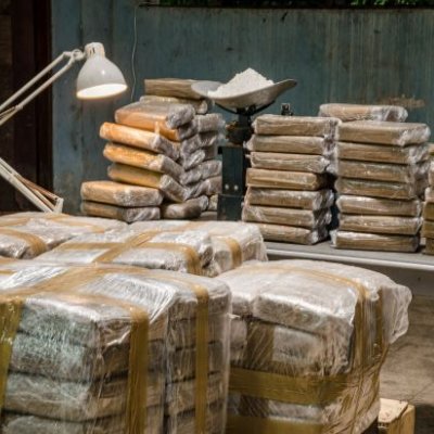 A warehouse with tables piled with illicit drug parcels and a desk light shining down.